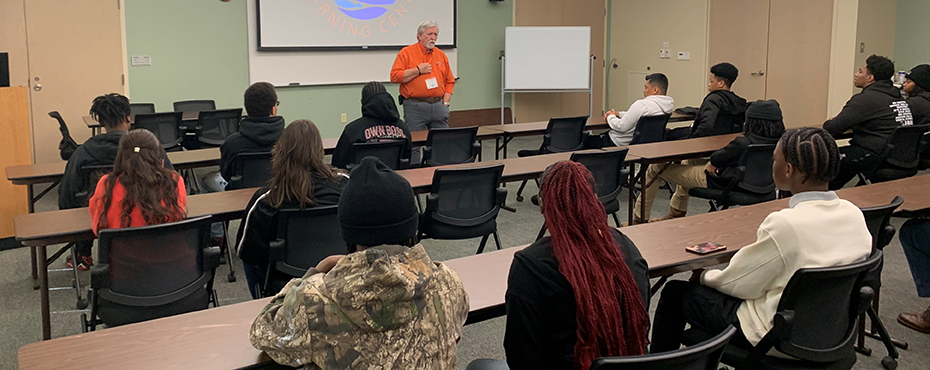 A male teacher presents a slideshow presentation to a group of students in a classroom setting