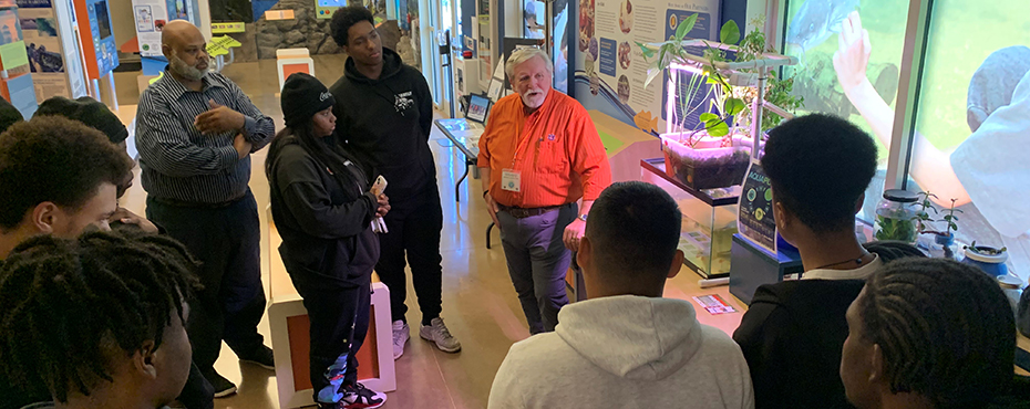 A male teacher presents an aquaponics lesson to a small group of students in a lab setting