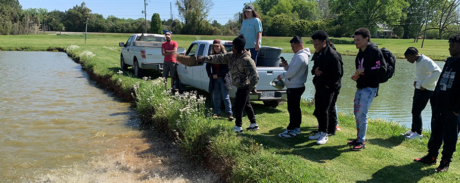 A male student tosses fish food to a pond for feeding while fellow students and adult teachers observe
