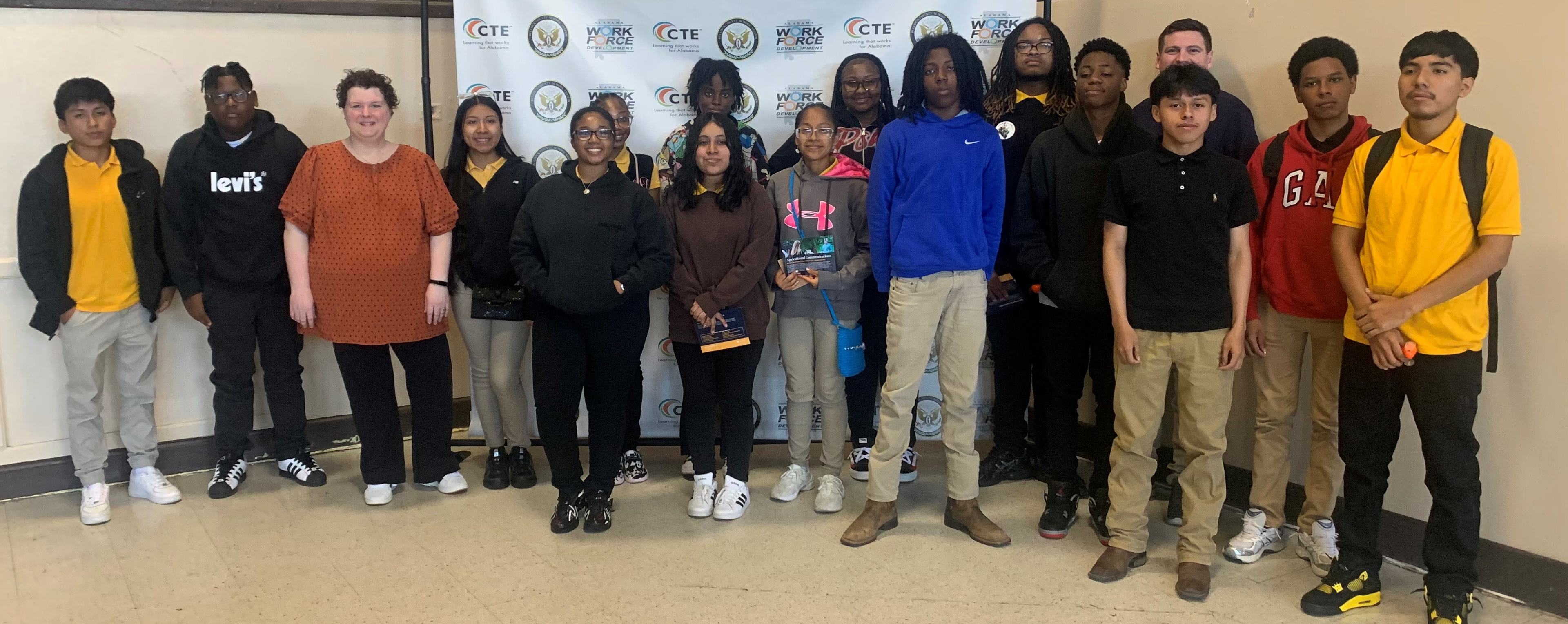 A large group of male and female students along with 2 adult presenters stand in front of a Career Technical Education backdrop in a school auditorium for a picture