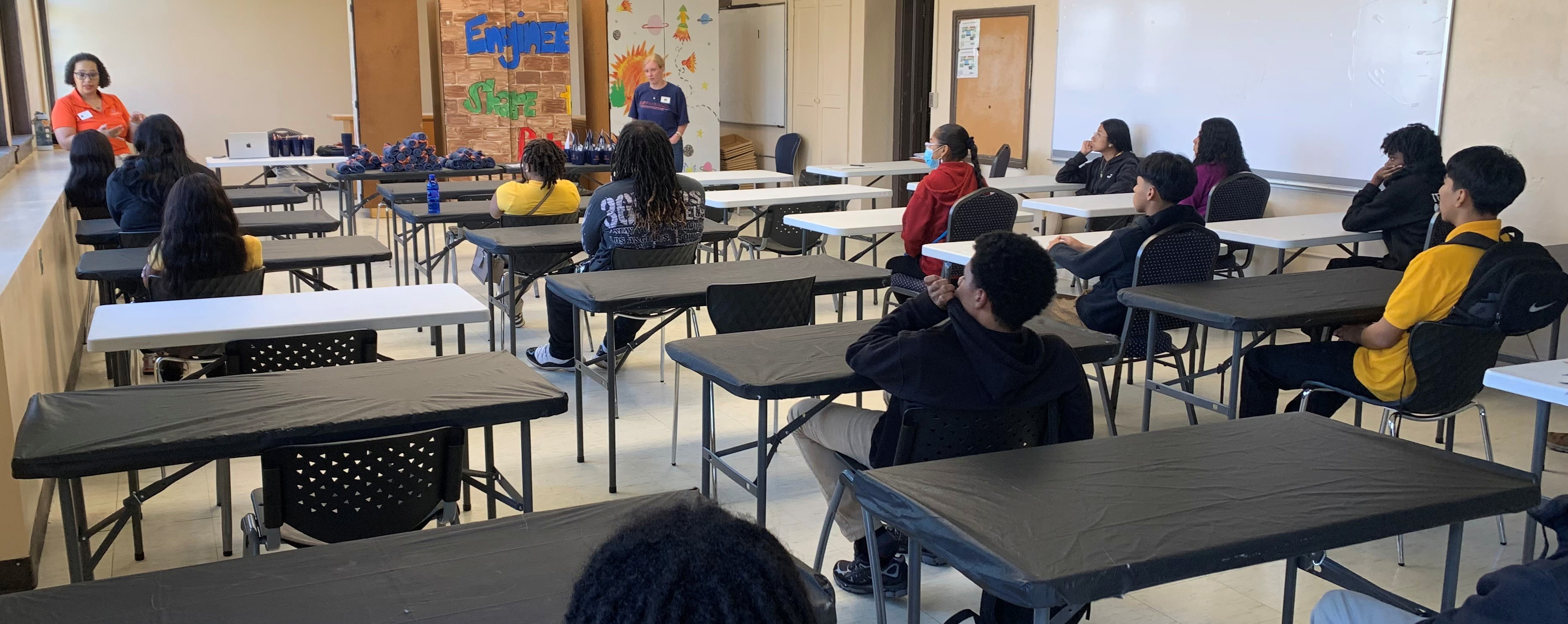 Two instructors stand in front of a classroom with students seated at tables.