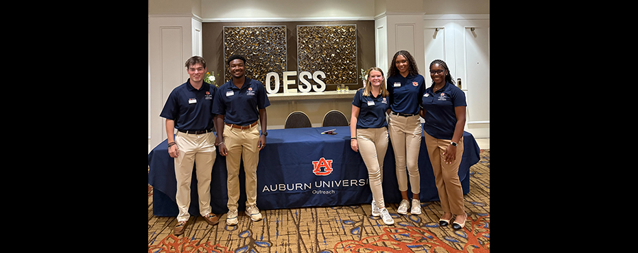 2 male and 3 female student workers stand for a picture at a registration table to welcome guests
