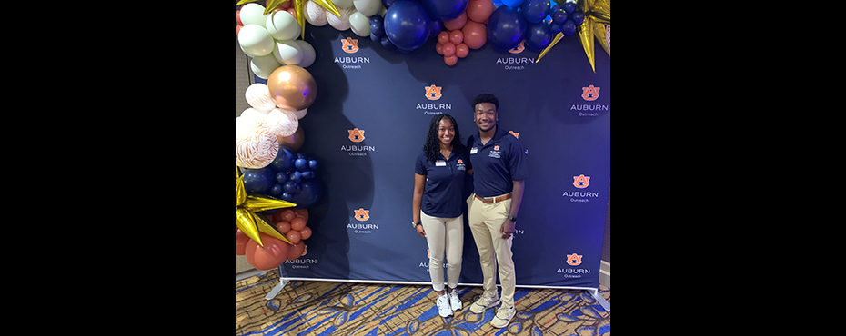 One female student worker and one male student worker stand for a picture with a University Outreach backdrop behind them