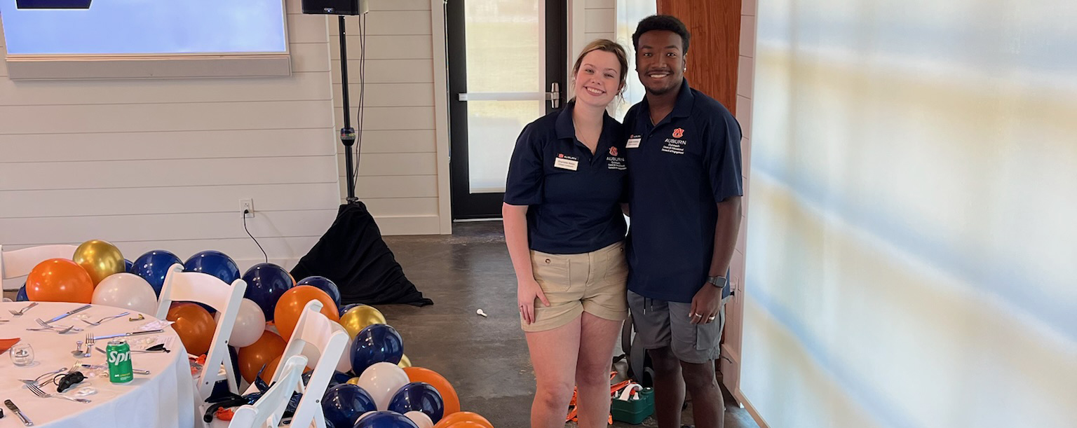 A female and male student pose for a picture in an event center while working to set up a balloon arc for the event
