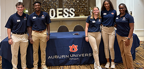 2 male and 3 female student workers stand for a picture at a registration table to welcome guests