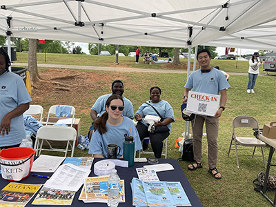 several volunteers sitting at a table outside, one standing holding a check-in sign