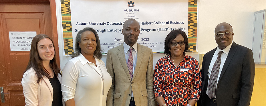 Three women and two men pose for a picture in front of a program banner.