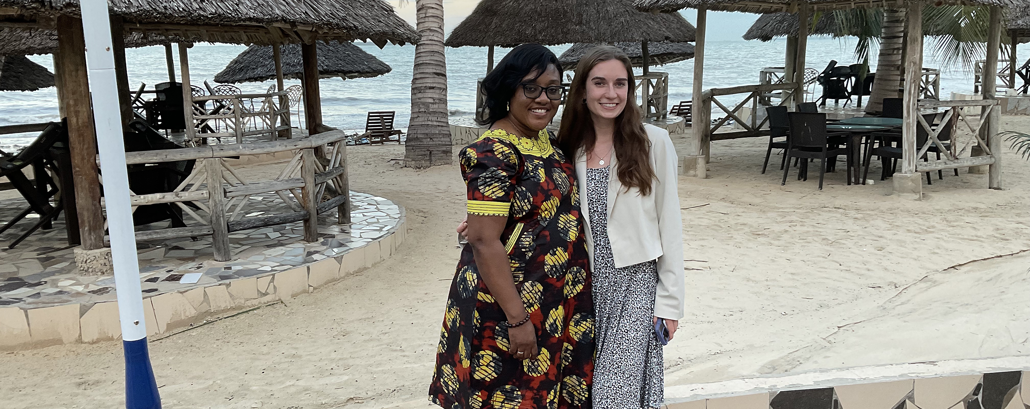 Two women standing by sand, trees, and huts.