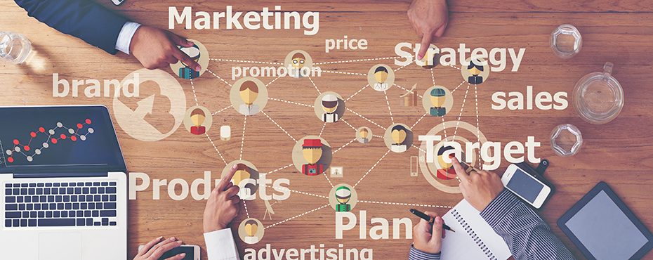 Hands and laptops at edge of table with words such as 'marketing' writting in the middle of the table.