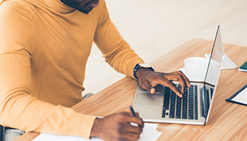 Black man with yellow shirt works on computer while writing on piece of paper