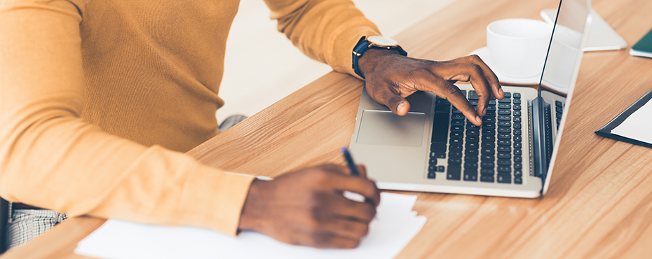 Black man in yellow shirt uses laptop and writes on a piece of paper.
