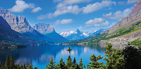 A tree-lined lake reflects the blue sky and the Rocky Mountains in the distance.