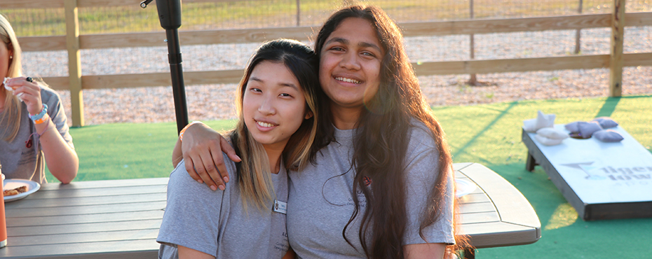 2 camp staff members smiling, outdoors with cornhole board in background
