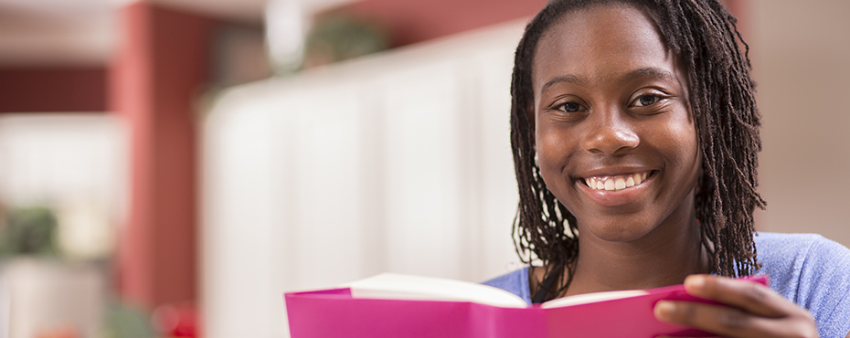 Young female sitting on coach and smiling at camera while holding a book with pink cover and wearing a purple shirt