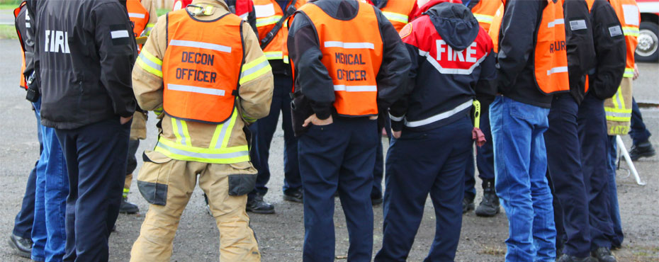 people wearing emergency uniforms, fire jackets, orange safety vests, all standing in a circle talking