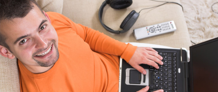 Man sitting on couch smiling up at camera with laptop on his laplap and headset and remote on couch.