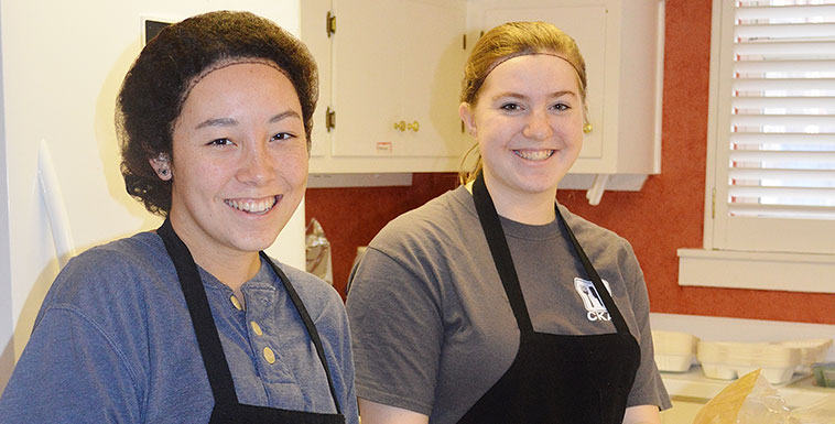 Two females smilling at camera wearing hair nets and aprons